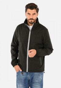 Giacca leggera nera con zip frontale, strisce bianche lungo la zip, polsini elastici e tasche laterali; abbinata a jeans blu.