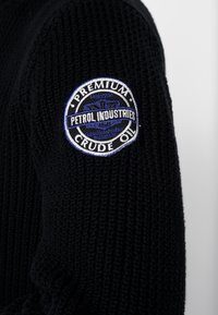 Czarny dzianinowy sweter z okrągłą naszywką z napisem "Petrol Industries" i "Premium Crude Oil" w kolorze niebieskim i białym. Teksturowana tkanina, trwały design.