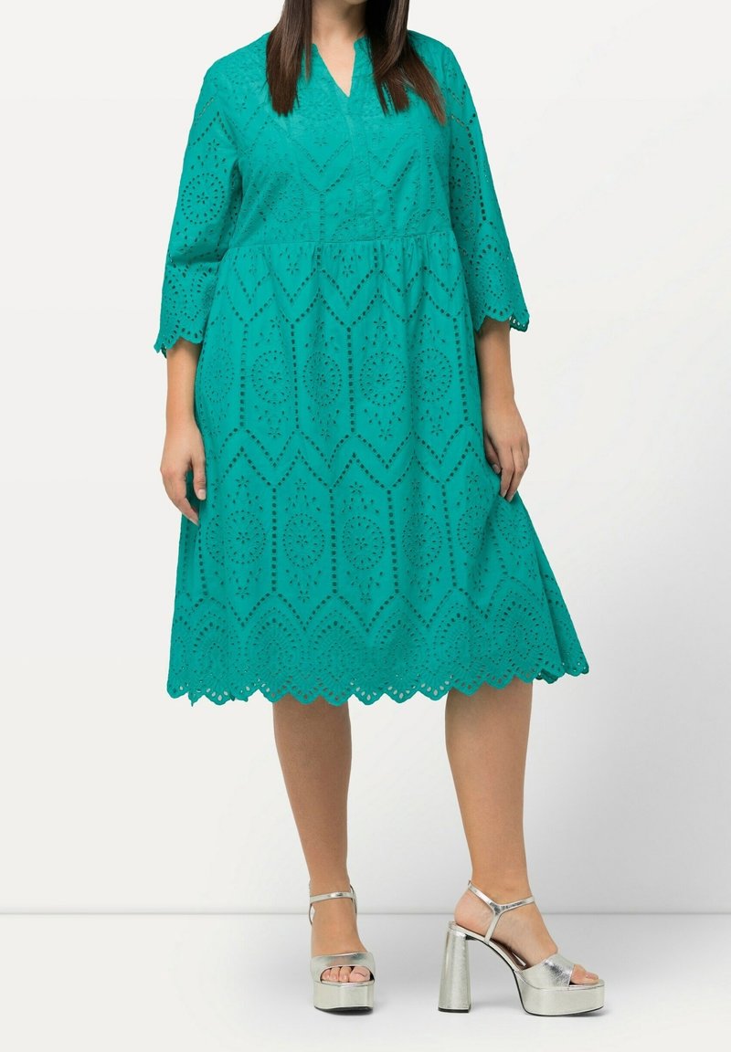 Femme portant une robe bleu sarcelle longueur genoux avec motifs œillets, manches trois-quarts, et des sandales plateforme à talons hauts argentées.