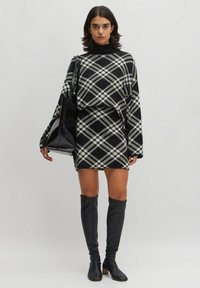 Zwart-wit geruite gebreide jurk met lange mouwen, hoge kraag en een getailleerd silhouet, gecombineerd met een zwarte glanzende tote bag en knielange laarzen.