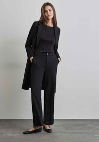 Blazer nero su un top nero luccicante, abbinato a pantaloni neri e ballerine. L'outfit presenta tasche e un dettaglio con bottoni sui pantaloni.