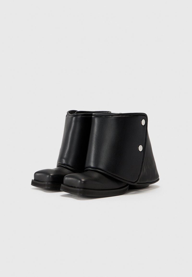 Zwarte enkellaarsjes van soepel leer, met een gevouwen ontwerp en drukknopdetails, en een ronde neus. Matte afwerking, platte zool.