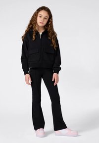 Camisola preta com fecho de correr e dois bolsos frontais, combinada com calças pretas ajustadas e ténis de cor rosa claro. Tecido liso, design casual.