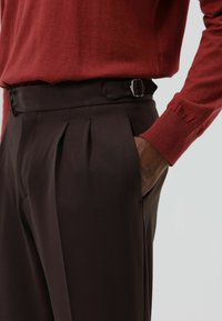 Bruine geplooide broek met een donkerriem en zilveren gesp, gecombineerd met een rood shirt met lange mouwen. Gevlekt stof en een getailleerde pasvorm zichtbaar.