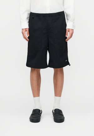 Bărbat purtând pantaloni scurți negri până la genunchi, cămașă albă cu mâneci lungi, șosete albe și pantofi slip-on negri, stând în fața unui fundal simplu.