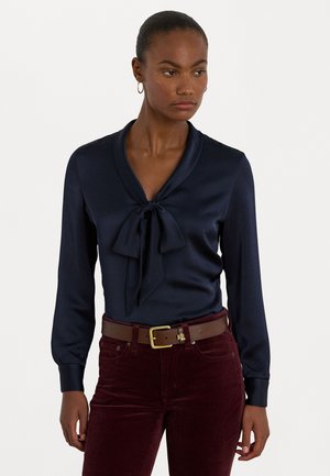 Marineblauwe satijnen blouse met een strikdetail bij de kraag, lange mouwen en een gladde textuur, gecombineerd met bordeauxkleurige corduroy broek en een bruine riem.