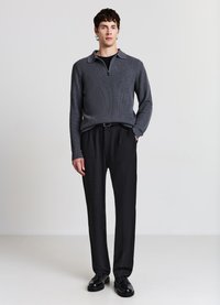 Grauer Strickpullover mit Reißverschlusskragen, kombiniert mit schwarzen, plissierten Hosen. Schwarze Schnürschuhe vervollständigen das Outfit.