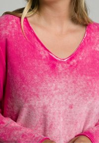 Růžový svetr s výstřihem do V, který má přechodový design, hladkou texturu a dlouhé rukávy s vroubkovanými manžetami. Látka působí měkce a lehce.