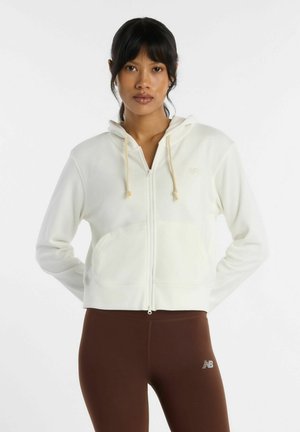 Donna con capelli scuri che indossa una felpa bianca con zip e leggings marroni, in piedi su uno sfondo bianco uniforme.