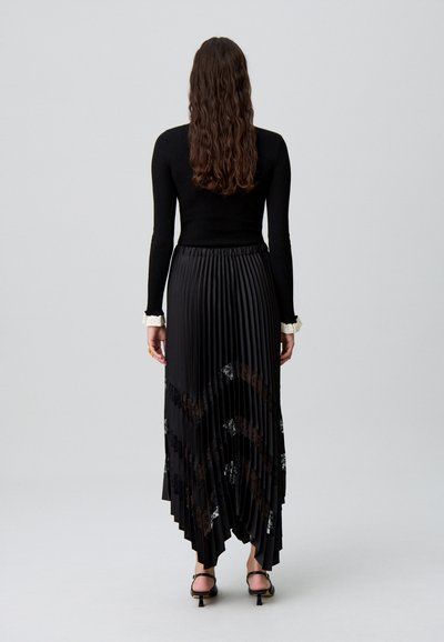 Jupe plissée noire avec des accents en dentelle, ceinture élastique et ourlet évasé. Associée à un haut ajusté noir à manches longues.