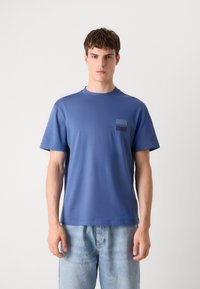 Zila kokvilnas t-krekls ar īsām piedurknēm, apaļu kakla izgriezumu un mazu logotipa uzliku kreisajā krūtī. Tam ir brīvs piegriezums un gluda tekstūra.