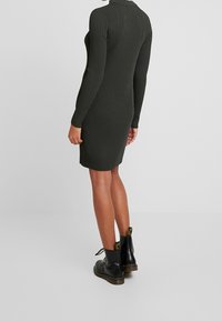 Robe en maille côtelée vert foncé avec manches longues, coupe ajustée et encolure ronde, associée à des bottines en cuir noir.