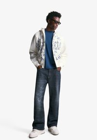 Weiße Zip-Hoodie mit blauer Schrift, blaues Rundhals-T-Shirt, dunkle Jeans und weiße Sneaker. Modell trägt Sonnenbrille.
