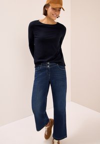 Mörkblå långärmad topp i marinblått som matchas med vida mörkblå jeans, med två knappar i gylfen, och bruna sneakers med vita snören.