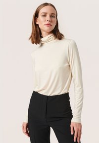 Kremasti turtleneck s dugim rukavima, mekanog materijala i širokog kroja, uparen s crnim hlačama visokog struka.