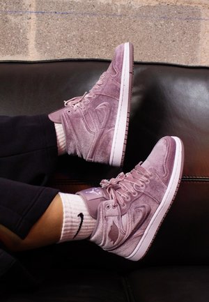 Voeten met lichtpaarse suède hoge sneakers en witte Nike-sokken, rustend op een zwarte leren bank tegen een grijze bakstenen muur.