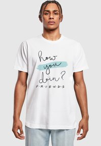 Camiseta de algodón blanca con el texto negro "¿cómo te va?" y "F.R.I.E.N.D.S." sobre un fondo de pinceladas azules. Mangas cortas, corte relajado.