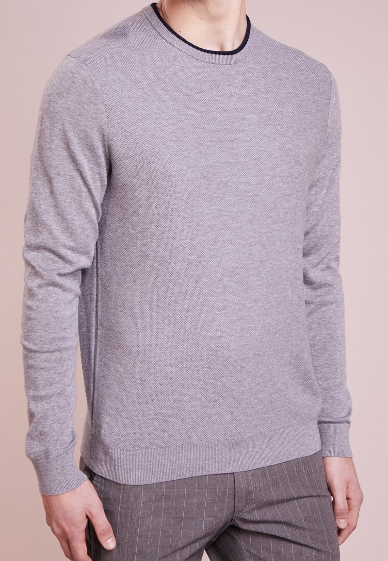 Homme portant un pull à manches longues gris clair texturé avec un pantalon gris foncé à fines rayures, sur un fond pâle.