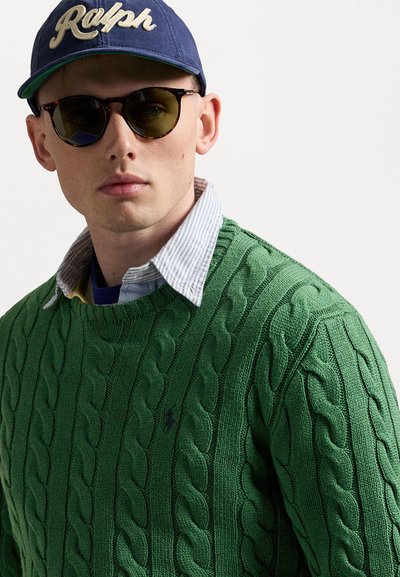 Green knitted sweater with cable patterns, layered over a striped collared shirt, paired with sunglasses and a blue cap featuring "Ralph" logo.