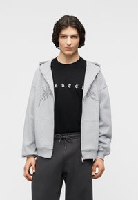 Νεαρό άτομο με μεσαίου μήκους μαλλιά, φορούν ανοιχτό γκρι φούτερ με φερμουάρ πάνω από μαύρο μπλουζάκι με στάμπα και σκούρο παντελόνι με κορδόνι.