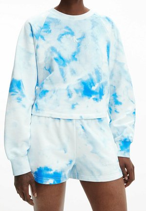 Femme portant un sweat-shirt à manches longues blanc et bleu tie-dye et un short assorti, avec de petits logos Calvin Klein sur la poitrine et le short.