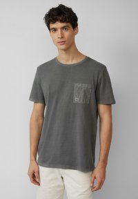 Grijze katoenen T-shirt met een ronde hals, korte mouwen en een patchzak op de linkerkant van de borst. Zachte textuur met minimale stikseldetails.