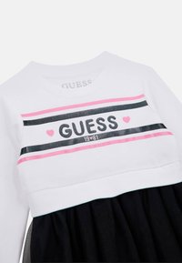 Vit sweatshirt-topp med svarta och rosa horisontella ränder; "GUESS" i fet svart text, parat med en svart kjol. Slät textur.