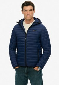 Mężczyzna w granatowej pikowanej kurtce z kapturem, noszący białą koszulę, z rękami w kieszeniach, na tle gładkiej białej ściany.