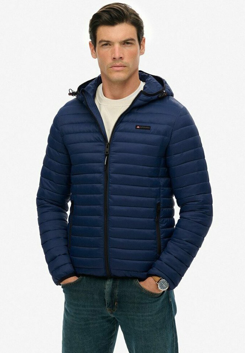 Mężczyzna w granatowej pikowanej kurtce z kapturem, noszący białą koszulę, z rękami w kieszeniach, na tle gładkiej białej ściany.