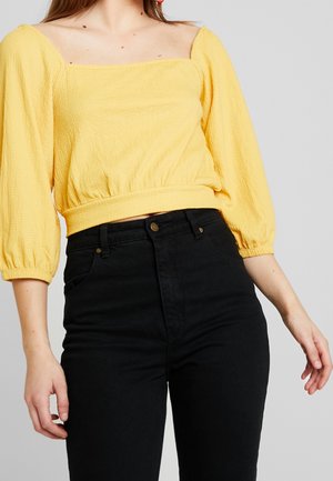 Gelber, strukturierter Oberteil mit quadratischem Ausschnitt und Puffärmeln, kombiniert mit hoch taillierter schwarzer Jeans mit Taschen und einem goldenen Knopf.