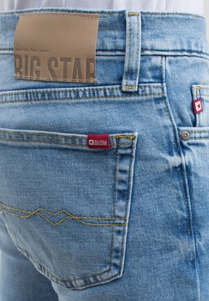 Jeans in denim azzurro con cuciture gialle, dotati di un'etichetta in pelle marrone del marchio e di un piccolo logo rosso sulla tasca posteriore.