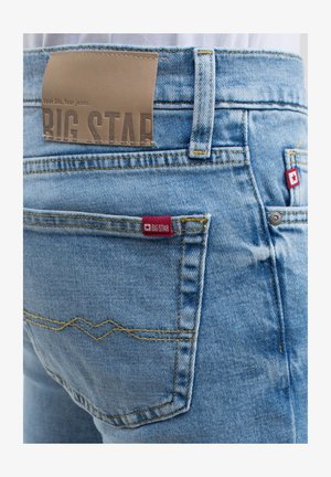 Világoskék farmernadrág sárga varrásokkal, barna bőr márkacímkével és egy kis piros logóval a hátsó zsebben.