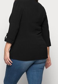 Zwarte tuniek met driekwart mouwen, voorzien van oprolmanchetten. Zacht, glad materiaal en licht getailleerde silhouet, gecombineerd met blauwe jeans.