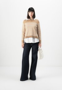 Beige-weißer gestreifter Strickpullover über einer weißen Bluse mit Kragen, kombiniert mit dunkelblauen weit geschnittenen Jeans und einer kleinen weißen Handtasche.