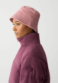 Lyserød kordfløjl bucket hat med bred skygge, båret med en højhalset lilla jakke med et rundt logo på ærmet.