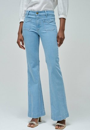 Femme portant un jean flare taille haute bleu clair avec poches avant et des sandales blanches à talons, debout devant un fond uni.