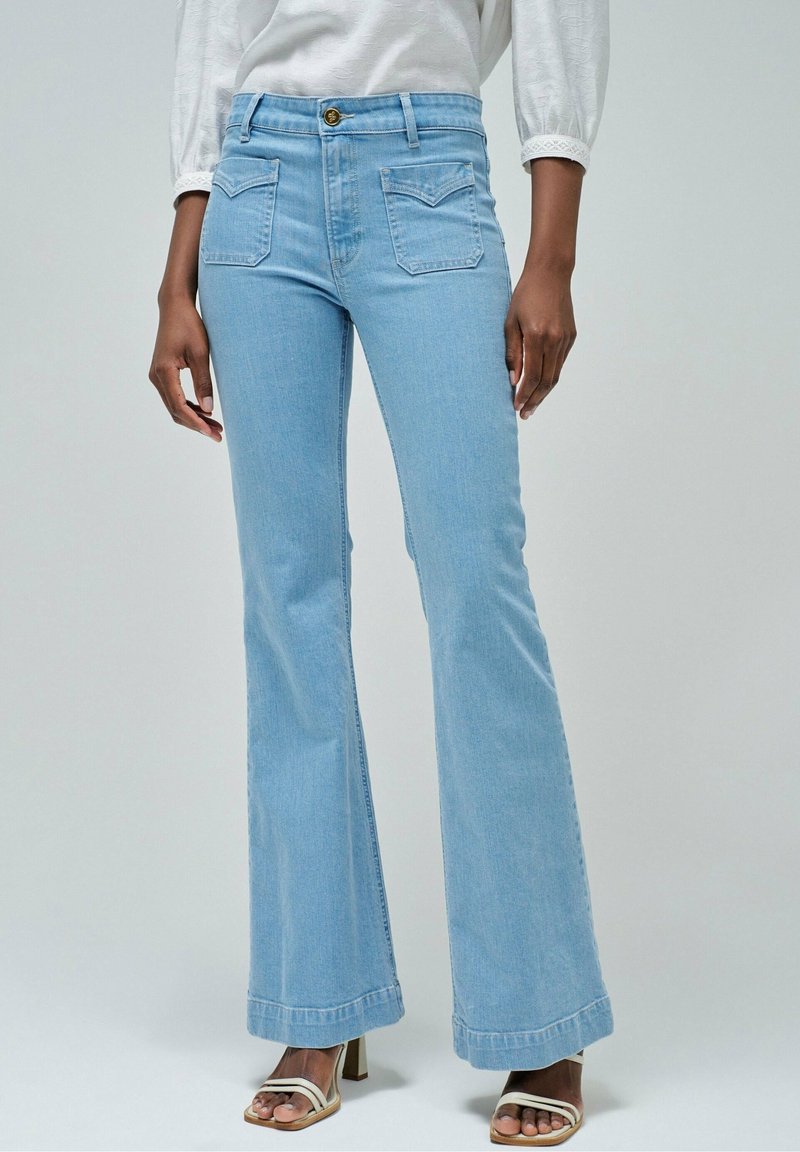 Femme portant un jean flare taille haute bleu clair avec poches avant et des sandales blanches à talons, debout devant un fond uni.