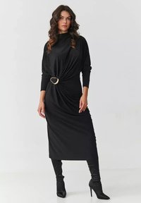 Czarna sukienka z długim rękawem, z przodu skręcona i ozdobiona złotym pierścieniem. Gładki materiał, długość do kostek, zestawiona z czarnymi kozakami za kolano.