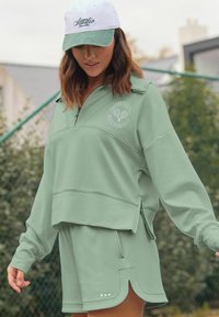 Costum sport verde deschis cu pulover cu fermoar până la piept și logo, fermoar lateral, și pantaloni scurți asortați, cu tiv curbat.