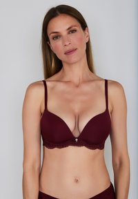 Vínová podprsenka s krajkovým lemováním, hlubokým výstřihem, hladkými košíčky, nastavitelnými ramínky a dekorativním stříbrným doplňkem uprostřed.