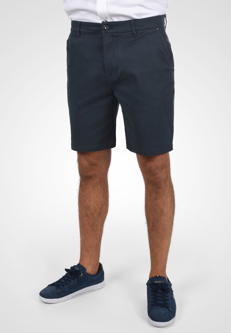 Shorts en coton bleu marine à coupe droite, fermeture à bouton unique et poches avant. Portés avec des baskets en suédine bleu marine assorties.