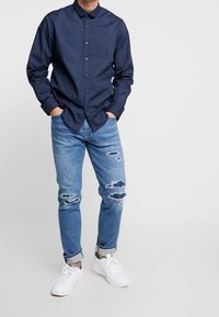Marinblå skjorta med krage, kombinerad med ljusblå slitna jeans med hål, och vita sneakers. Modern avslappnad outfit.
