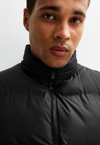 Czarna puchowa kurtka z wysokim kołnierzem, z zapięciem na zamek i gładką, strukturalną teksturą. Zbliżenie górnej części ciała.