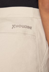 Béžové látkové šortky so vyšívaným logom "HOUDINI". Materiál má hladkú štruktúru s viditeľným šitím a vreckami.