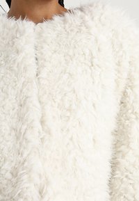 Hvid, fluffy jakke med et tekstureret, kunstigt pelsmateriale og en høj krave. Har ingen synlige lukninger eller mønstre.