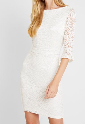 Biała koronkowa sukienka do kolan z przezroczystymi rękawami do łokcia i kwiatowym wzorem, noszona przez osobę o jasnej karnacji z długimi blond włosami.