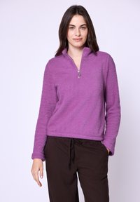 Pullover in pile viola con colletto alto e dettaglio con zip, caratterizzato da un tessuto testurizzato, abbinato a pantaloni marroni a righe.