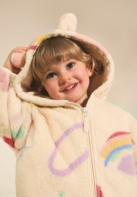 Sweat à capuche en polaire couleur crème avec motifs arc-en-ciel, oreilles d'ours et fermeture éclair. Texture douce et design ludique avec des accents colorés.