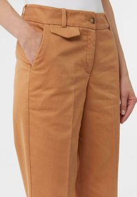 Pantaloni di cotone beige con taglio dritto, dotati di tasche laterali, una piccola tasca a soffietto e un dettaglio in ottone alla vita.