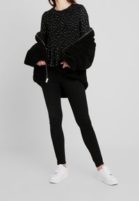 Black, textured oversized coat with a zipper, worn over a black top with white polka dots and paired with black fitted jeans and white sneakers.